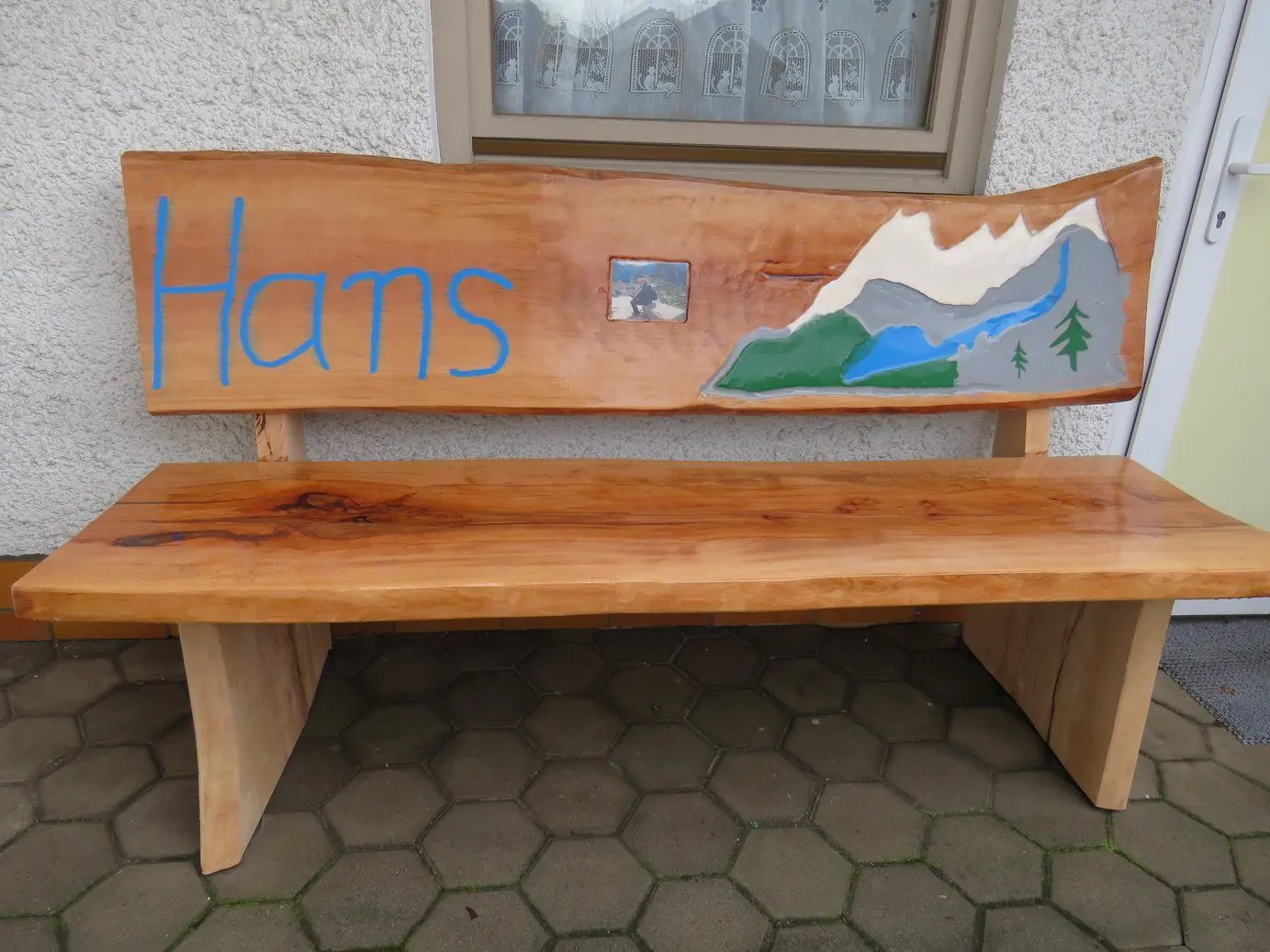 Holzbank mit eingeschnitztem Namen und gemaltem Bergmotiv vor einem Haus